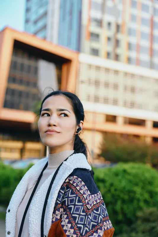 Woman outdoor city settings wearing cardigan, gazing upward with thoughtful look, showcasing fashion and street style amid daylight, architecture, greenery and urban ambiance