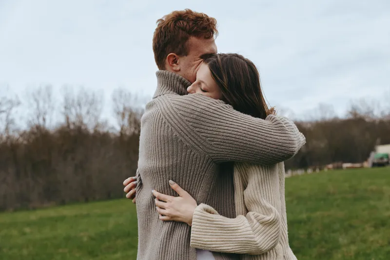 Young couple in cozy knit sweaters embraces warmly outdoors on a cloudy day in a green field, expressing love and comfort in a natural lifestyle setting.