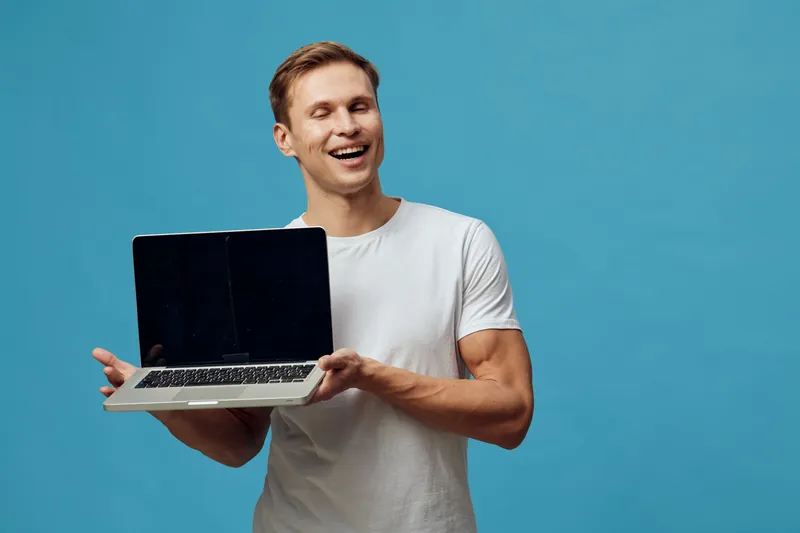 Smiling young man with light brown hair, wearing casual white t-shirt, holding open laptop computer, looking cheerful and confident, isolated on plain blue background, studio portrait. People lifestyl