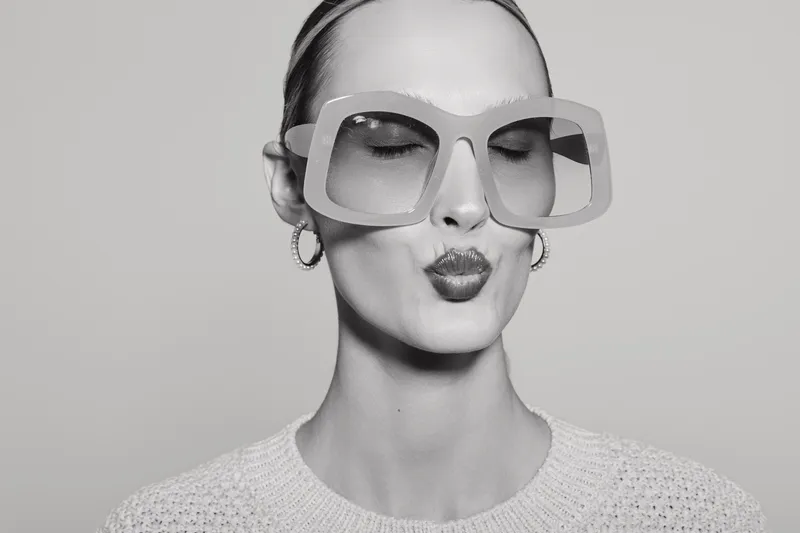 Young white woman with pale skin wearing oversized sunglasses and hoop earrings makes a playful kiss face on a plain background. Concept of fashion, fun, and youth.