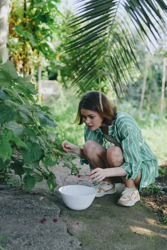 Woman crouches in a lush garden outdoors, picking fruit into a white bowl. Bright natural light, calm mood, and close connection with plants convey harvest, care, nourishment, and simple rural life.