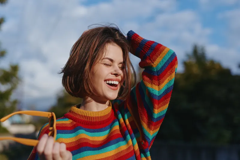 Woman smiling in colorful striped sweater, short hair and casual outfit outdoors, joyful expression, holding sunglasses and bag strap, lifestyle portrait ideal for fashion or travel.