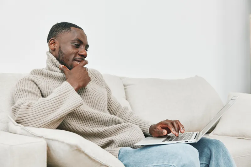 Thoughtful young man in a cozy sweater working on laptop in a bright living room, reflecting a modern lifestyle and creativity