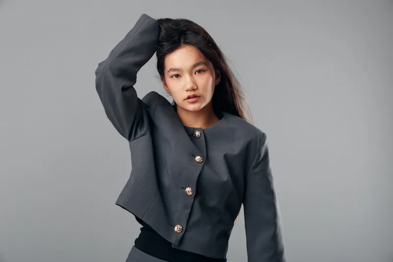 Young woman with long dark hair wearing a buttoned gray jacket posing against plain background with hand raised to head showcasing thoughtful expression and modern style.