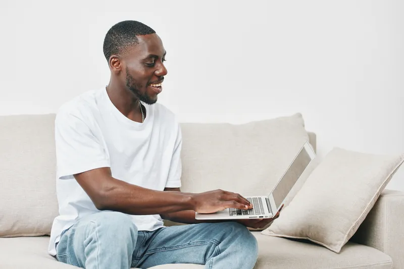 Smiling Black man working on a laptop at home, dressed casually in a white t shirt, conveying happiness and productivity in a bright, minimalist living room setting