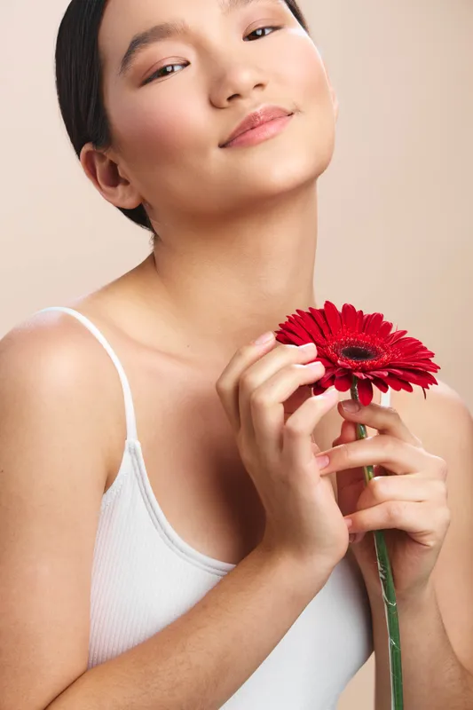 Asian woman smiles softly as she holds a vivid red flower close to her chest, creating a graceful studio portrait with warm lighting and a serene, confident mood.