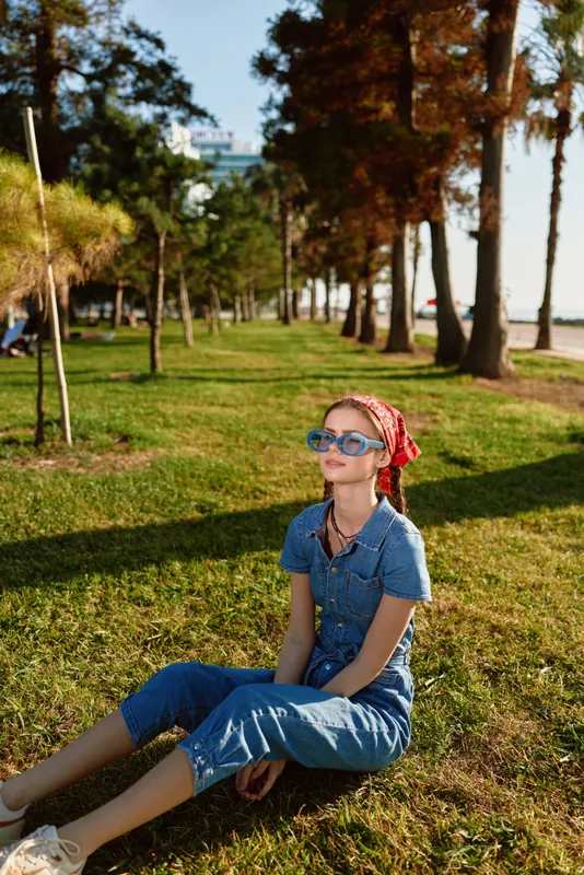 lifestyle woman wearing modern boho-western denim street style outfit with sunglasses and red bandana sitting on grass under sunlight in warm film color tones