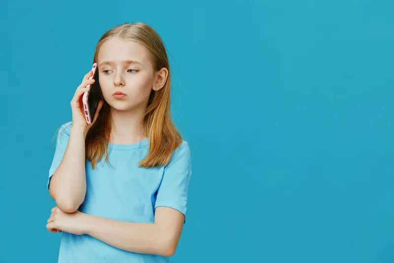 Sad girl in a blue shirt talking on a smartphone, against a bright blue background, expressing emotions of surprise and disappointment, ideal for family and teenage themes