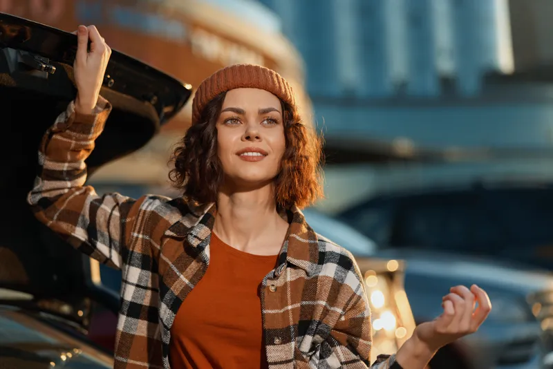 Woman by car trunk with beanie and flannel shirt, smile and curly hair in urban golden hour glow, authentic candid lifestyle portrait conveying mindful living, emotional storytelling and authenticity.