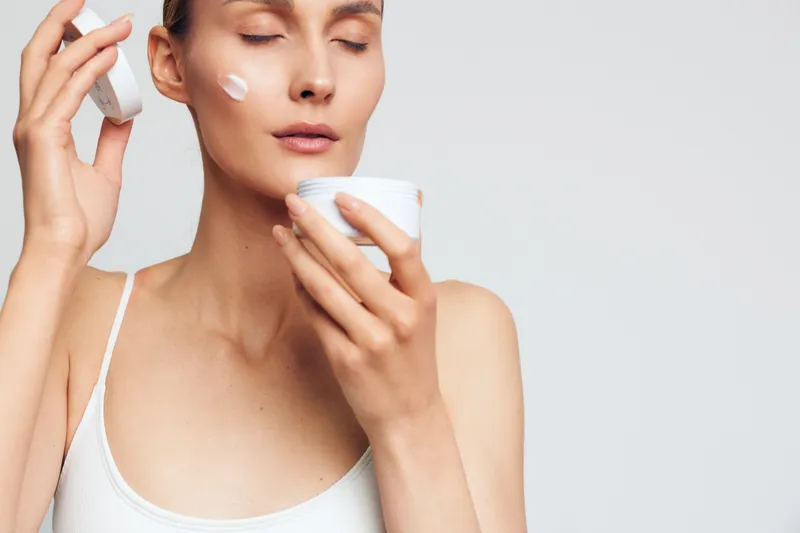 Young woman with brunette hair applies face cream with closed eyes in a bright minimalistic room wearing a white tank top, focusing on skincare and self-care routine.