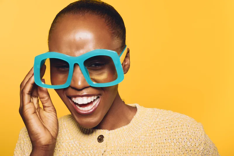Happy African woman with dark skin wearing oversized blue glasses and a beige sweater smiles brightly against yellow background. Concept of joy, fashion, and vibrant portrait.