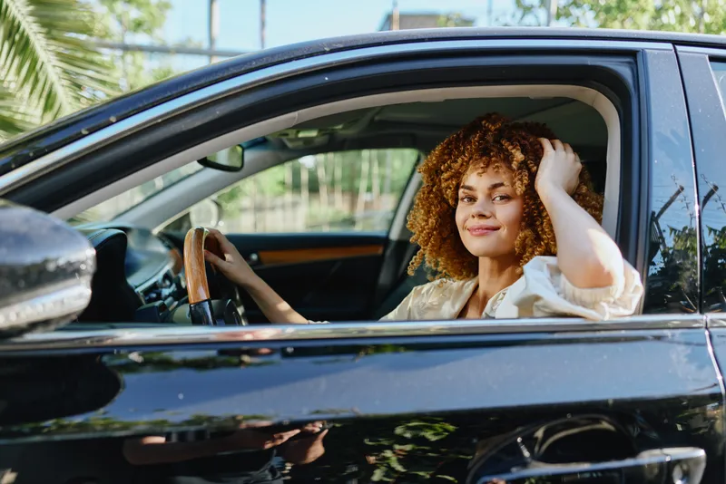 smiling woman with curly hair driving black car outdoors in sunny weather, relaxed and confident young African American female behind steering wheel
