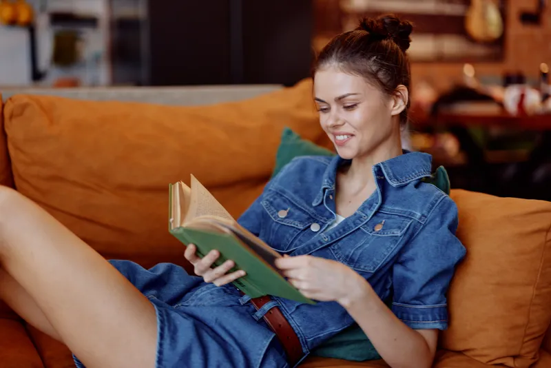 Женщина в джинсовом платье читает книгу, сидя на диване в интерьере кофейни