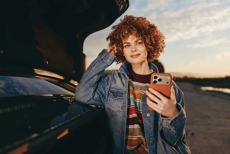 Woman lifestyle at sunset by car, curly hair denim jacket, holding smartphone for a casual selfie, one hand touching face, relaxed smile, golden hour outdoor road background