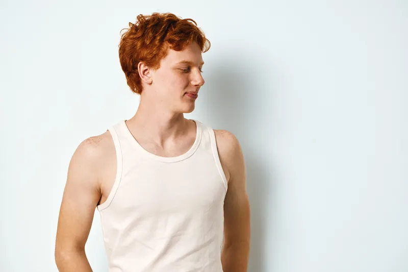 Young man with red hair wearing white tank top standing against white wall looking sideways with neutral expression in casual clothing and natural light portrait.