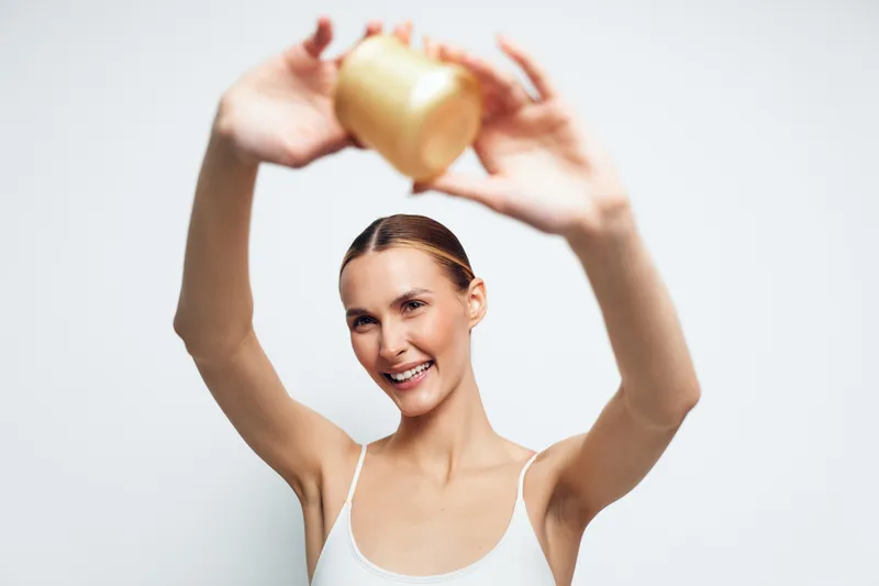 A happy young woman with slicked back hair holds a jar of cream above her head smiling in a bright studio. Concept for skincare, beauty routine, and natural cosmetics.