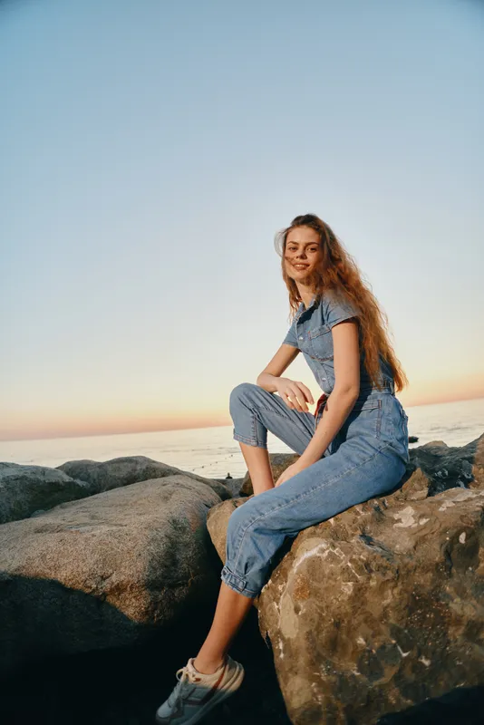 Lifestyle woman in modern boho-western denim street style sitting on rocks at sunset with relaxed smile and natural light in film color tone.