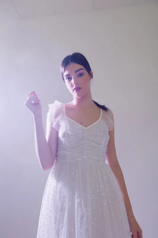Young woman in a delicate white dress poses against a soft light background. Her expression is calm and elegant, highlighting beauty and fashion concepts.