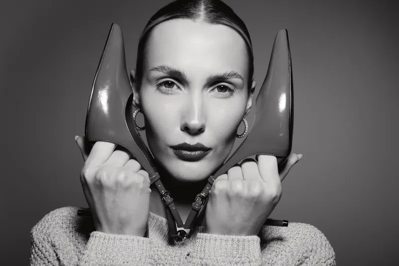 Young white woman with pale skin holds two high heel shoes near face in black and white portrait. Concept for fashion, beauty, style, and minimalism in studio.