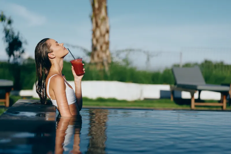Young woman in white swimsuit relaxing in outdoor pool drinking a red beverage through a straw. Sunny day with clear sky and green background. Concept of summer lifestyle and leisure.