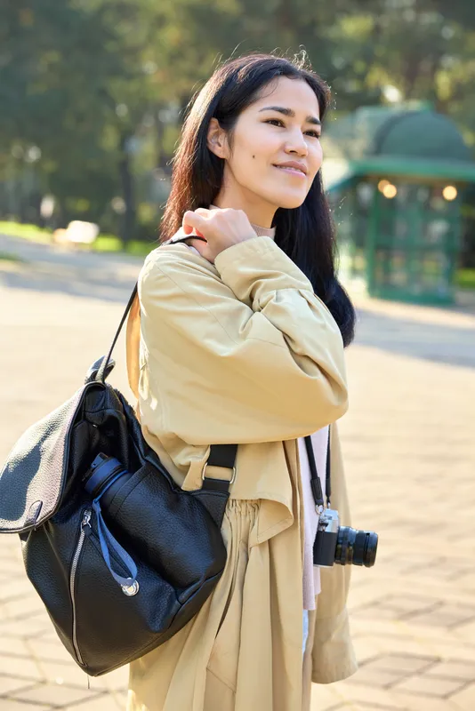 Outdoor scene with a woman holding a camera, wearing a beige coat and carrying a backpack, ready for travel and adventure in a bright sunny park, calm and stylish