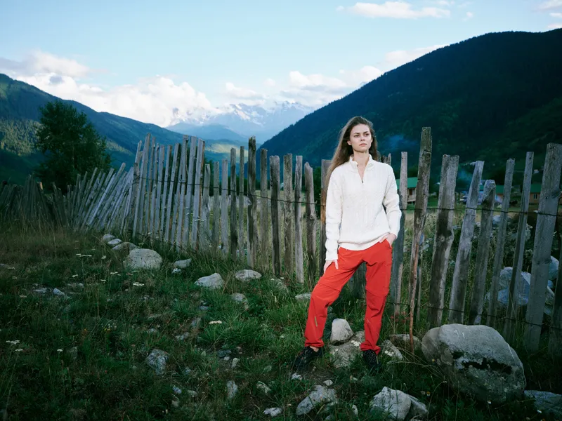 Outdoor mountain landscape with a woman in a white sweater and red pants standing beside a wooden fence on rocky terrain under a bright blue sky and distant snowy peaks