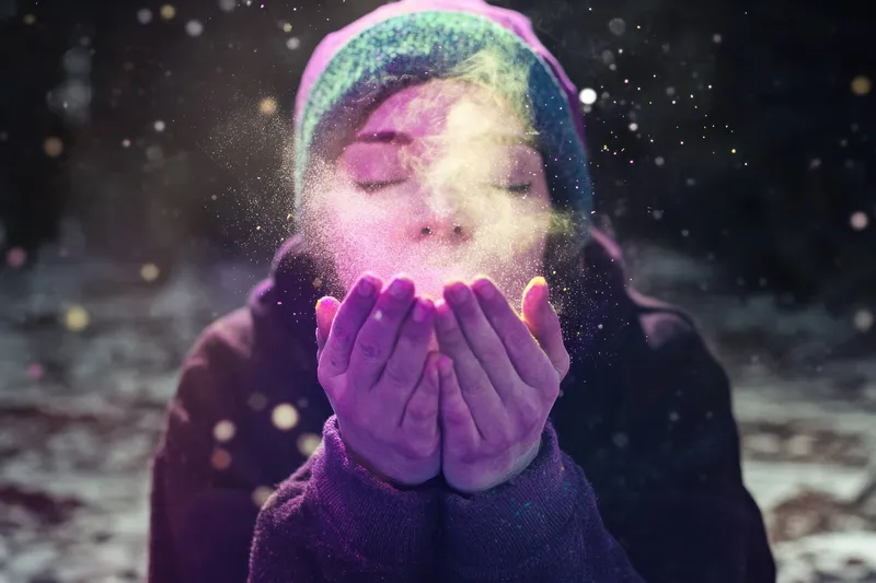 Молодая женщина с радостным выражением лица раздувает разноцветную пудру в заснеженном лесу, создавая волшебную атмосферу