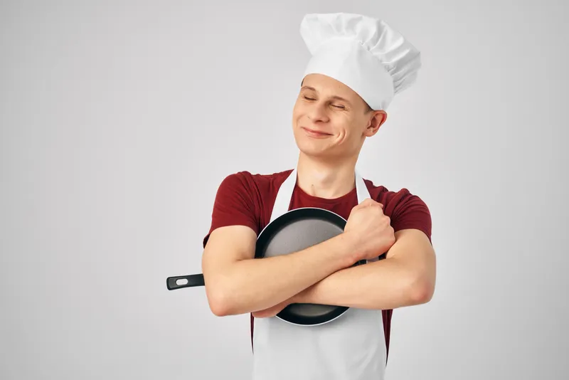 Мужчина в поварской форме держит в руках сковороду на профессиональной кухне