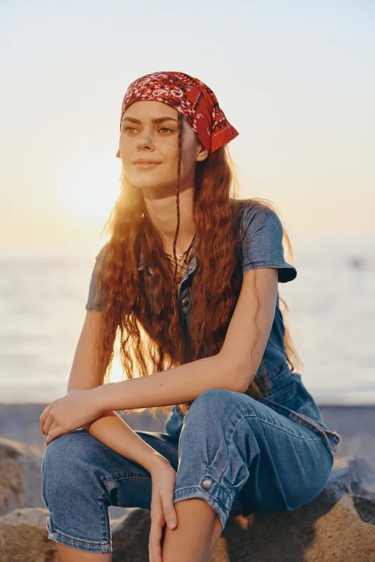lifestyle woman in modern boho-western denim street style wearing red bandana sits relaxed by the beach with warm film color sunlight glowing behind her