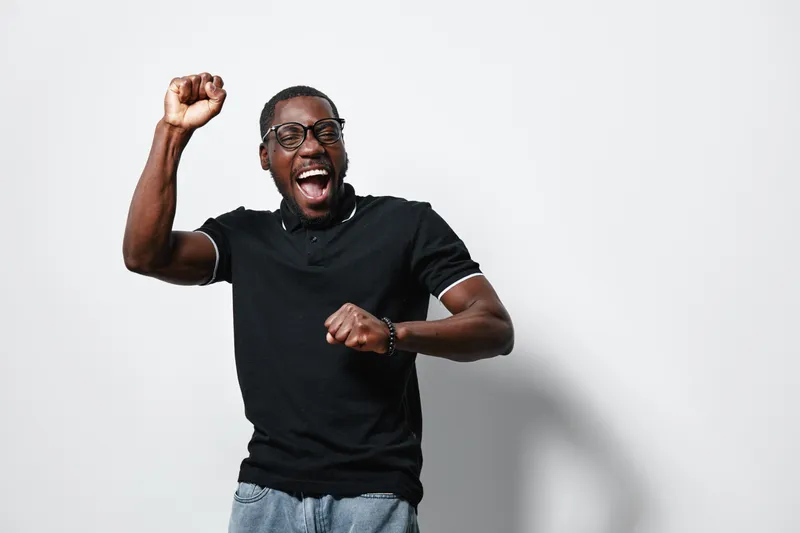 African American man with dark skin wearing glasses and black polo shirt celebrating with raised fist and joyful expression on white background. Casual style and happiness.