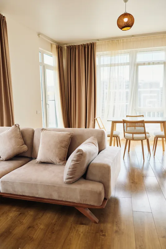 Sofa pillows curtains table floor living home bright interior with modern beige couch and cozy cushions, large windows with sheer drapes and warm hardwood floors in open plan apartment
