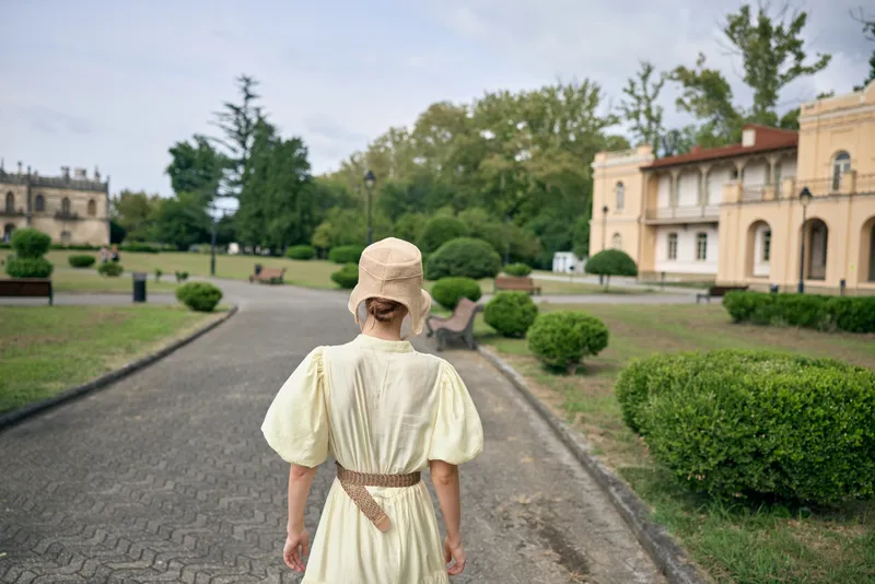 Young girl in a cream vintage dress walks along a gravel path in an expansive historic estate garden, creating a nostalgic and tranquil scene.