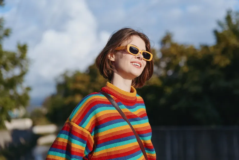 Young woman (20s) in a colorful striped sweater and yellow sunglasses smiling outdoors with short hair and crossbody bag, looking up in a sunny urban park lifestyle and travel portrait.