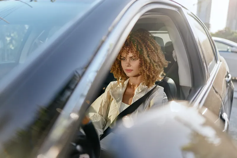 Woman driving car with serious expression wearing beige jacket and seatbelt in urban daylight setting, showing concentration and safety during daily commute.