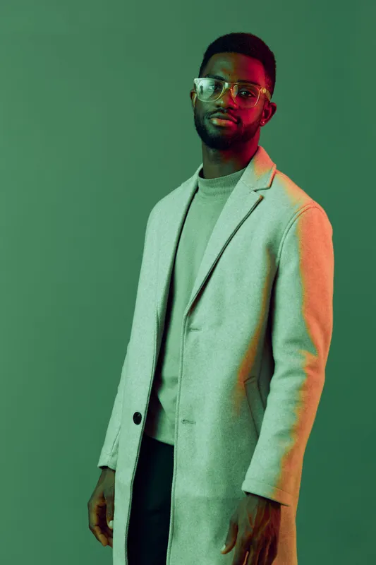 Stylish man with glasses wearing a light grey coat poses confidently against a vibrant green background, showcasing modern fashion and self assurance