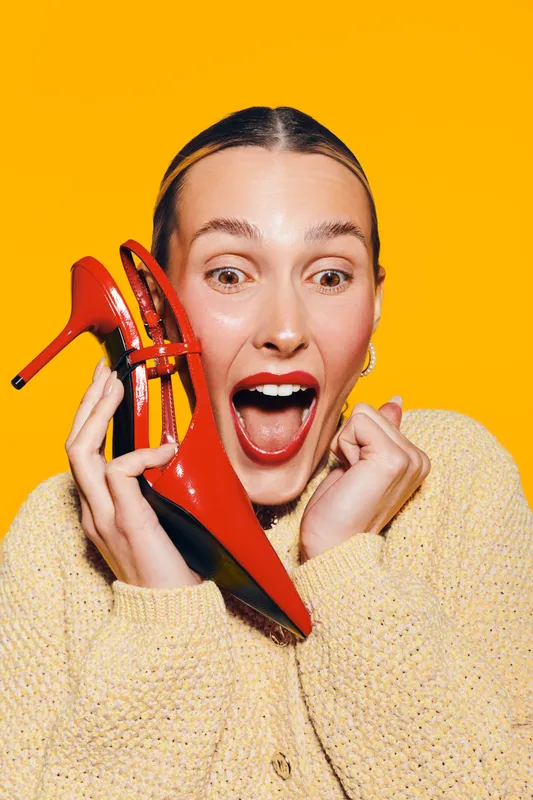 Excited woman in yellow sweater holds red high heel shoe near face with open mouth on bright yellow background. Concept of fashion, style, happiness, and shopping enthusiasm.