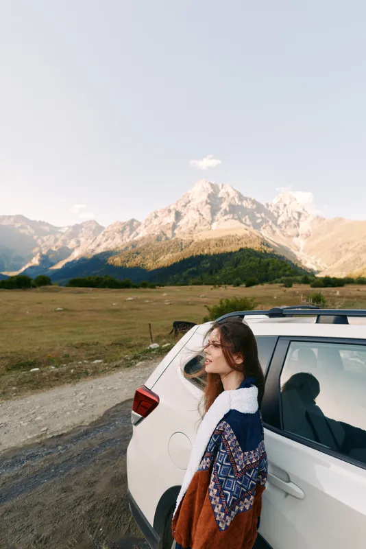 Woman and car parked by mountains on a travel roadtrip, landscape portrait in nature with autumn sweater, scenic meadow and sunlight creating calm adventure mood and freedom.
