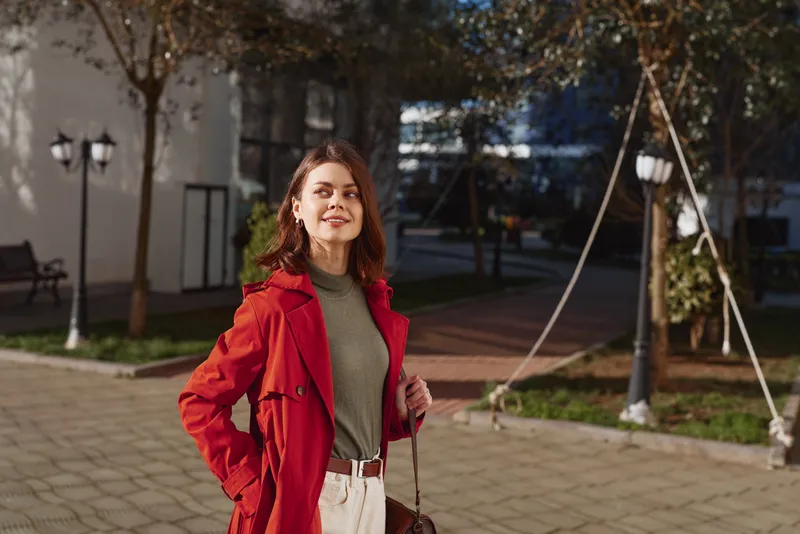 Woman outdoors, smiling, wearing a red coat over a green sweater, standing in a sunlit park with a casual stance, soft breeze, natural colors, leafy backdrop, warm light on fabrics