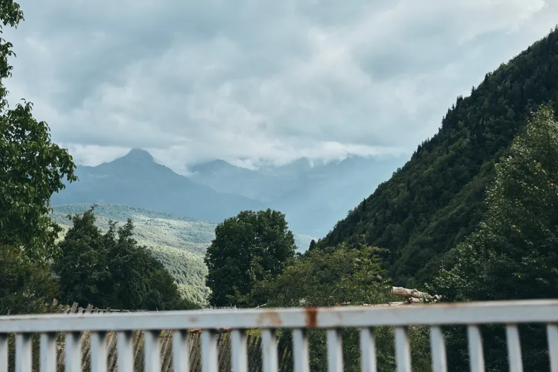 A tranquil mountain landscape with lush trees and distant peaks seen beyond a weathered railing, capturing calm natural scenery, open air, and an expansive sky.