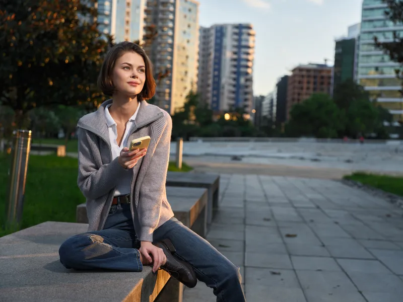 A relaxed young woman sits on a concrete ledge in a city park, gazing with a gentle smile while holding a smartphone, suggesting casual connection and urban style.