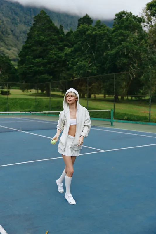 Stylish young female tennis player in a white sporty outfit on a blue tennis court, exuding confidence and joy while holding a tennis ball, with a lush green background