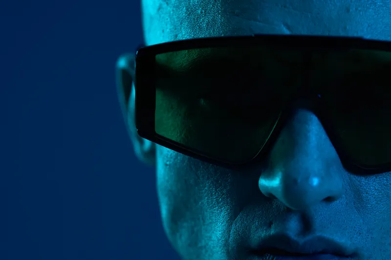Close-up of a man wearing futuristic sunglasses, showcasing a blue-toned atmosphere and striking facial features. The image conveys a sense of mystery and modernity.