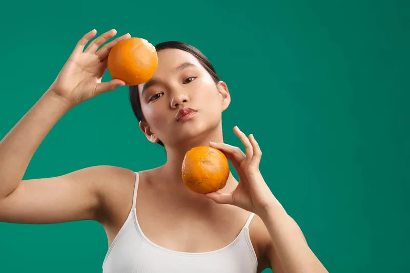 Asian woman poses with oranges in a vibrant studio, projecting playful confidence. Bright lighting and a clean backdrop highlight simple style and modern composition, inviting fresh energy and