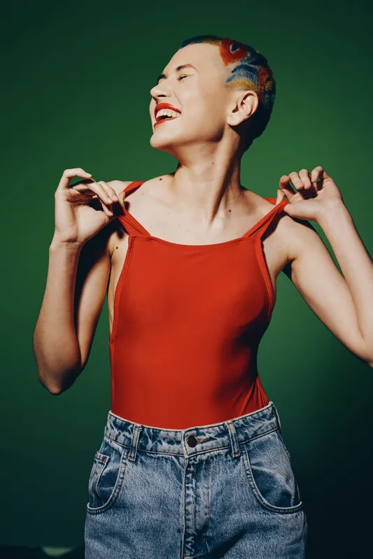 joyful woman with colorful short hair wearing a red tank top and denim jeans smiling brightly against a green backdrop in casual style