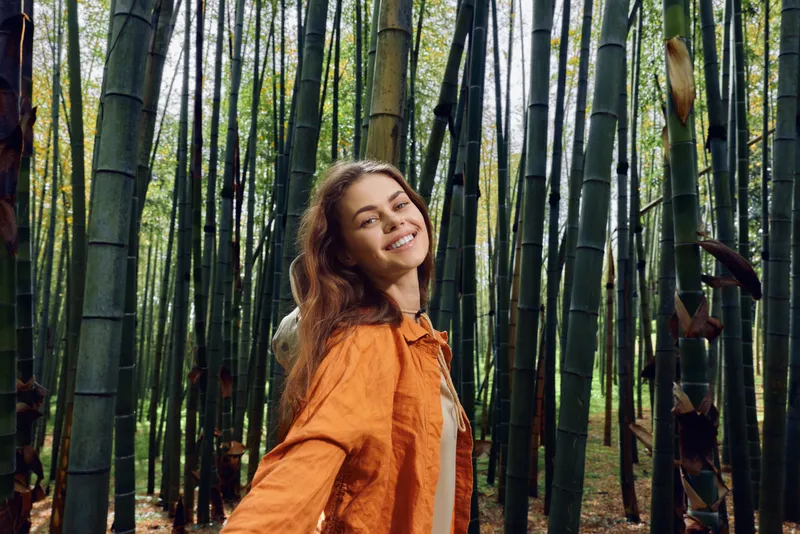 Woman bamboo forest portrait smiling and looking back, casual orange jacket photography moment, nature scene with tall green trees and dappled sunlight, joyful outdoor lifestyle travel.