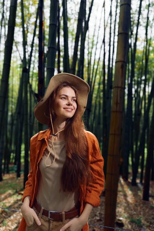 Woman with hat and long hair smiling in bamboo forest, portrait of a happy traveler in nature. Warm sunlight, casual orange jacket and relaxed pose among tall green stalks and trees.