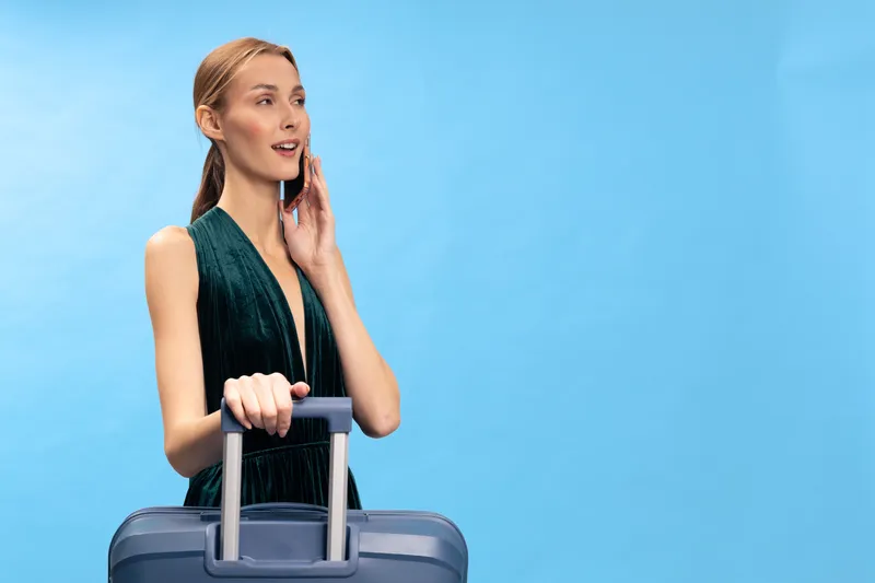 Young woman in green dress talking on phone while holding suitcase handle, blue background. Concept of travel, communication, and vacation preparation.
