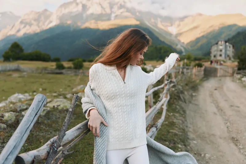 A woman in a white knit sweater stands along a wooden fence in a rural landscape, embracing the breeze for a calm outdoor lifestyle moment in the mountains.