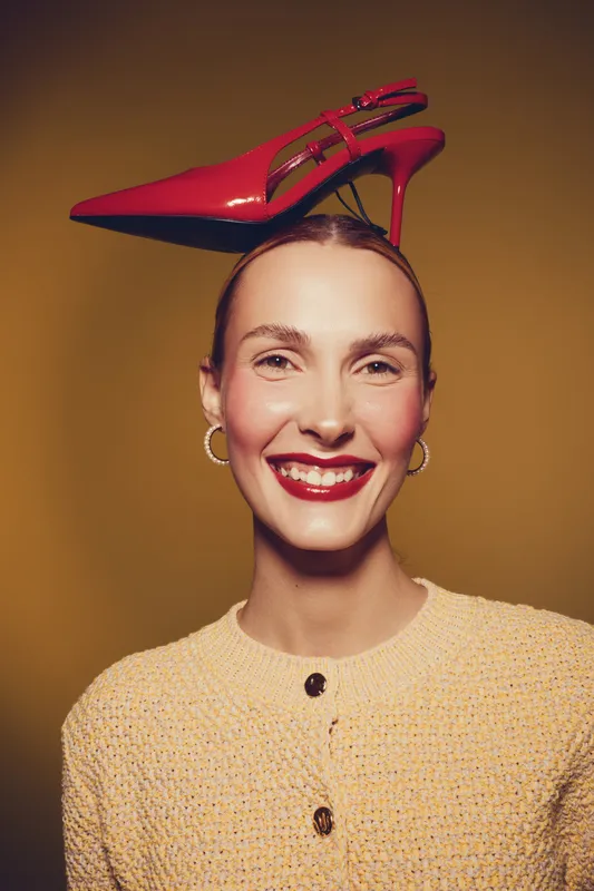 Smiling woman with red lipstick and gold earrings balances a red high heel shoe on her head against a warm brown background. Concept of fashion, style, and playful elegance.