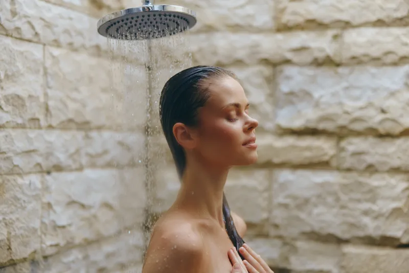 Woman enjoying outdoor shower with closed eyes, wet hair, natural stone wall background, relaxing atmosphere, skin care and wellness concept.